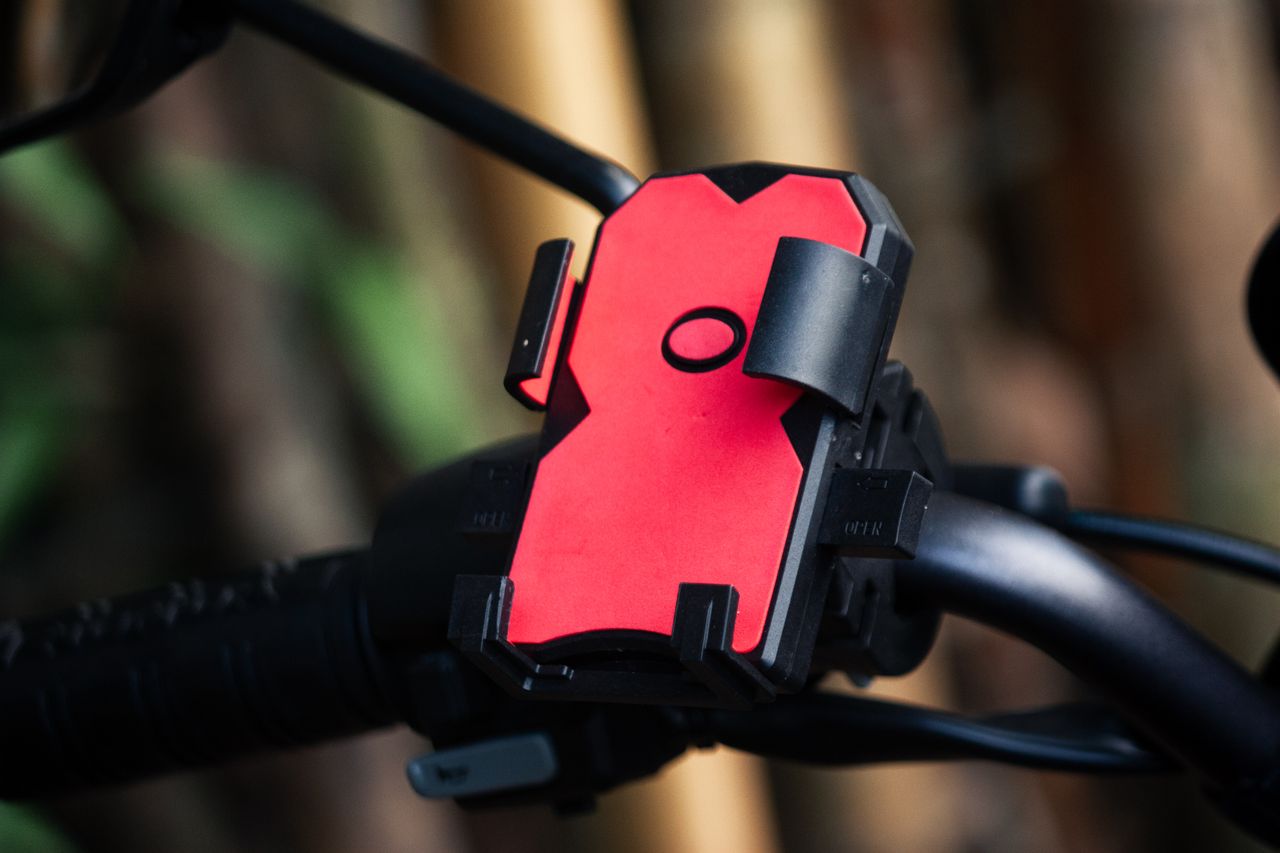 about-01 Close-up of a red phone holder mounted on a bicycle handlebar outdoors.